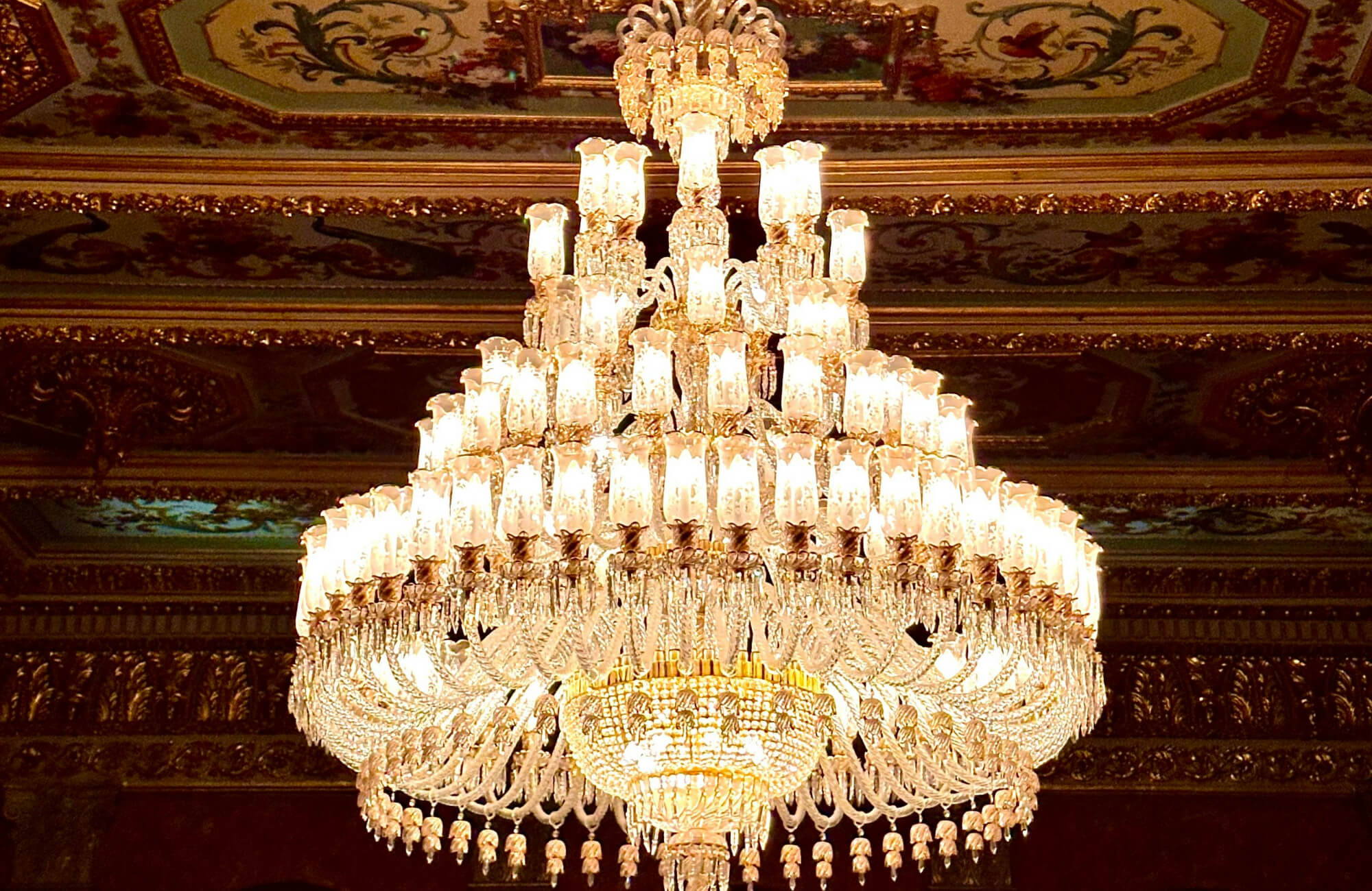 Opulent crystal chandelier with cascading tiers of glowing lights, creating a majestic focal point against the ornate, gilded ceiling.