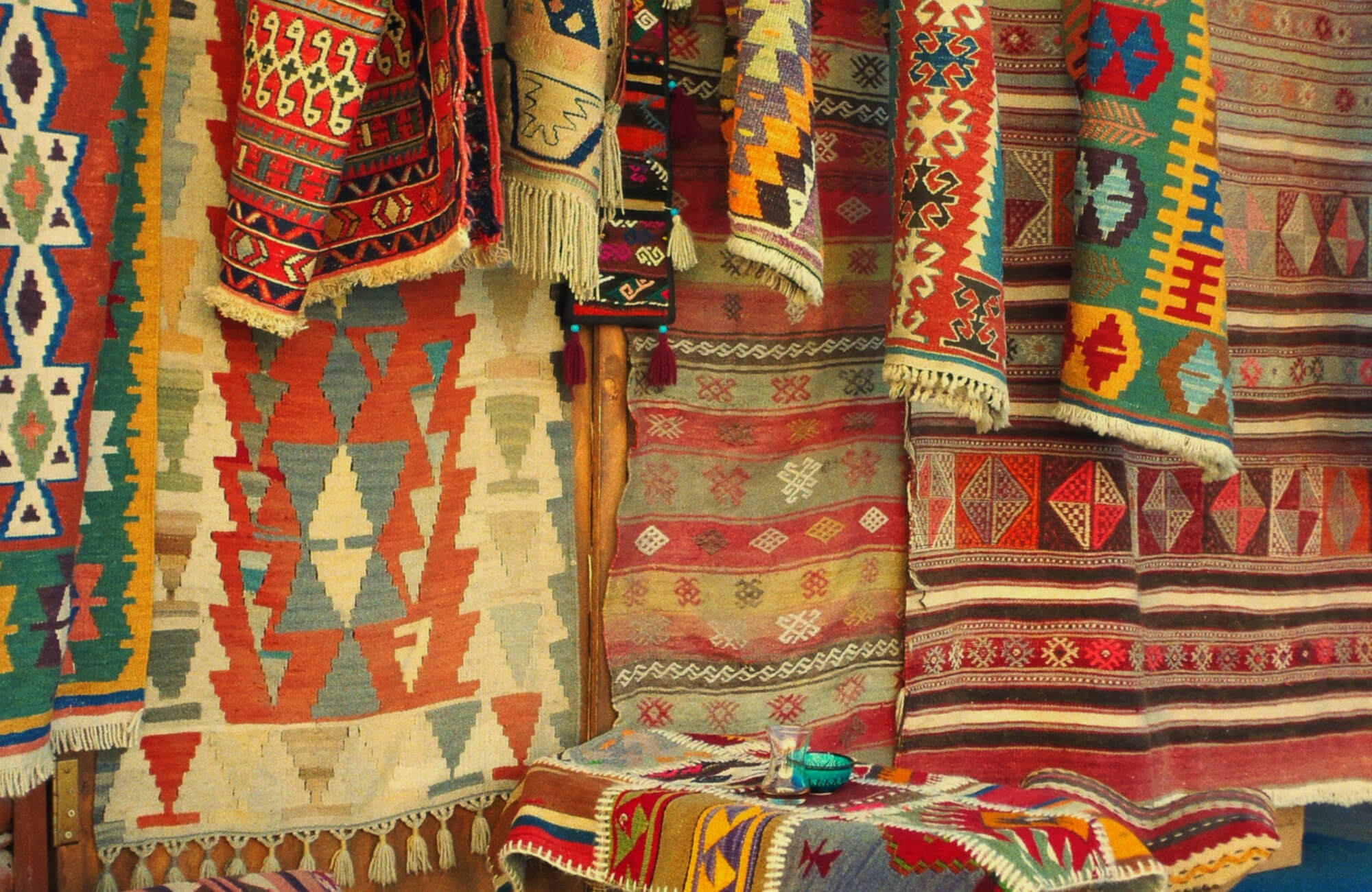 Colorful handcrafted rugs with tribal and geometric patterns displayed in layers at a traditional market stall.