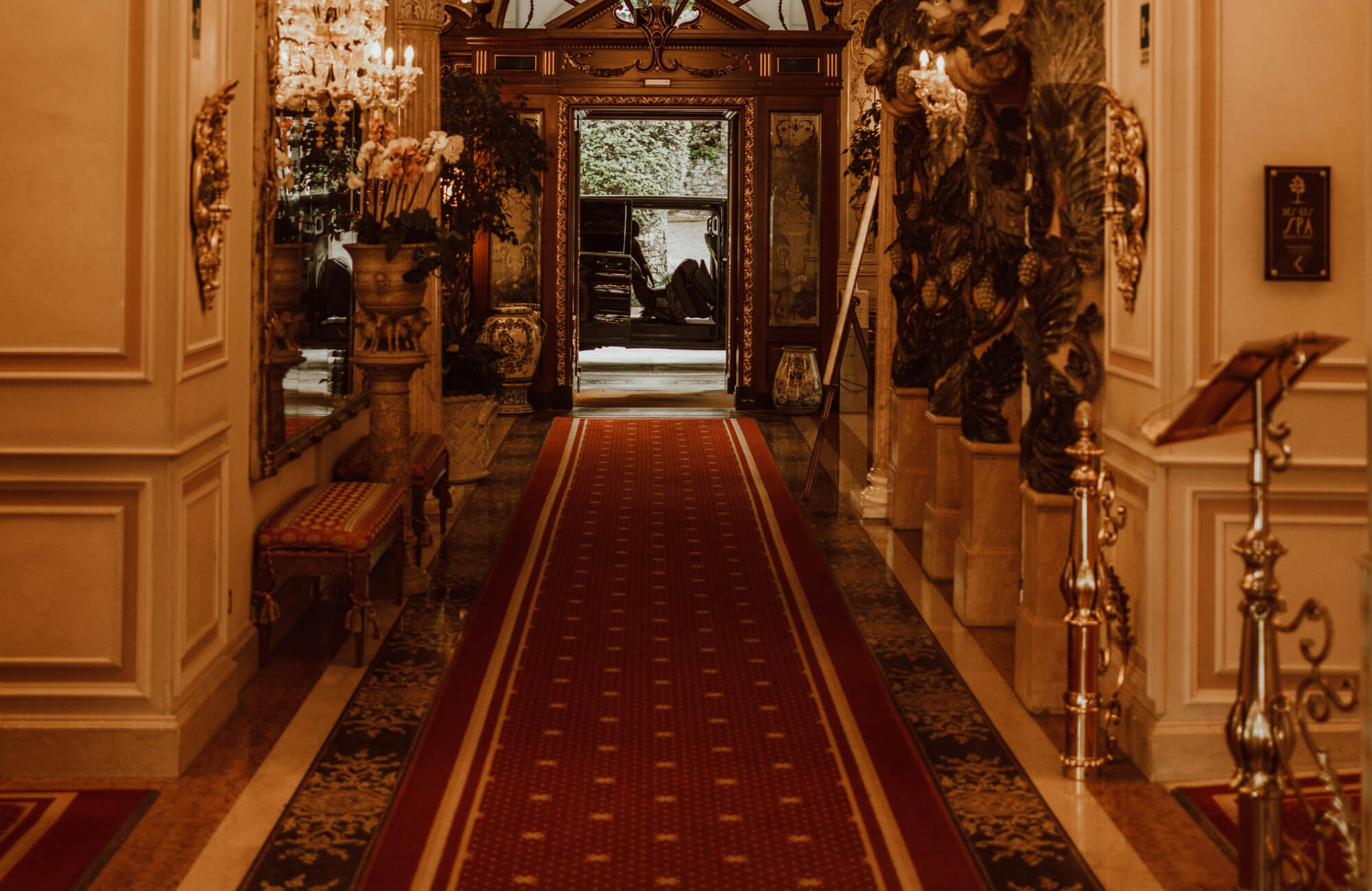 An opulent hallway features a long oriental rug runner in deep red with subtle geometric detailing, guiding the eye through gilded columns, crystal chandeliers, and richly ornamented walls that evoke classic European luxury.