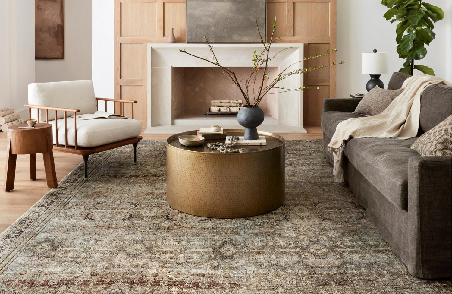 Living room with a patterned rug framed by a round brass table, styled around a fireplace, and a modern accent chair.
