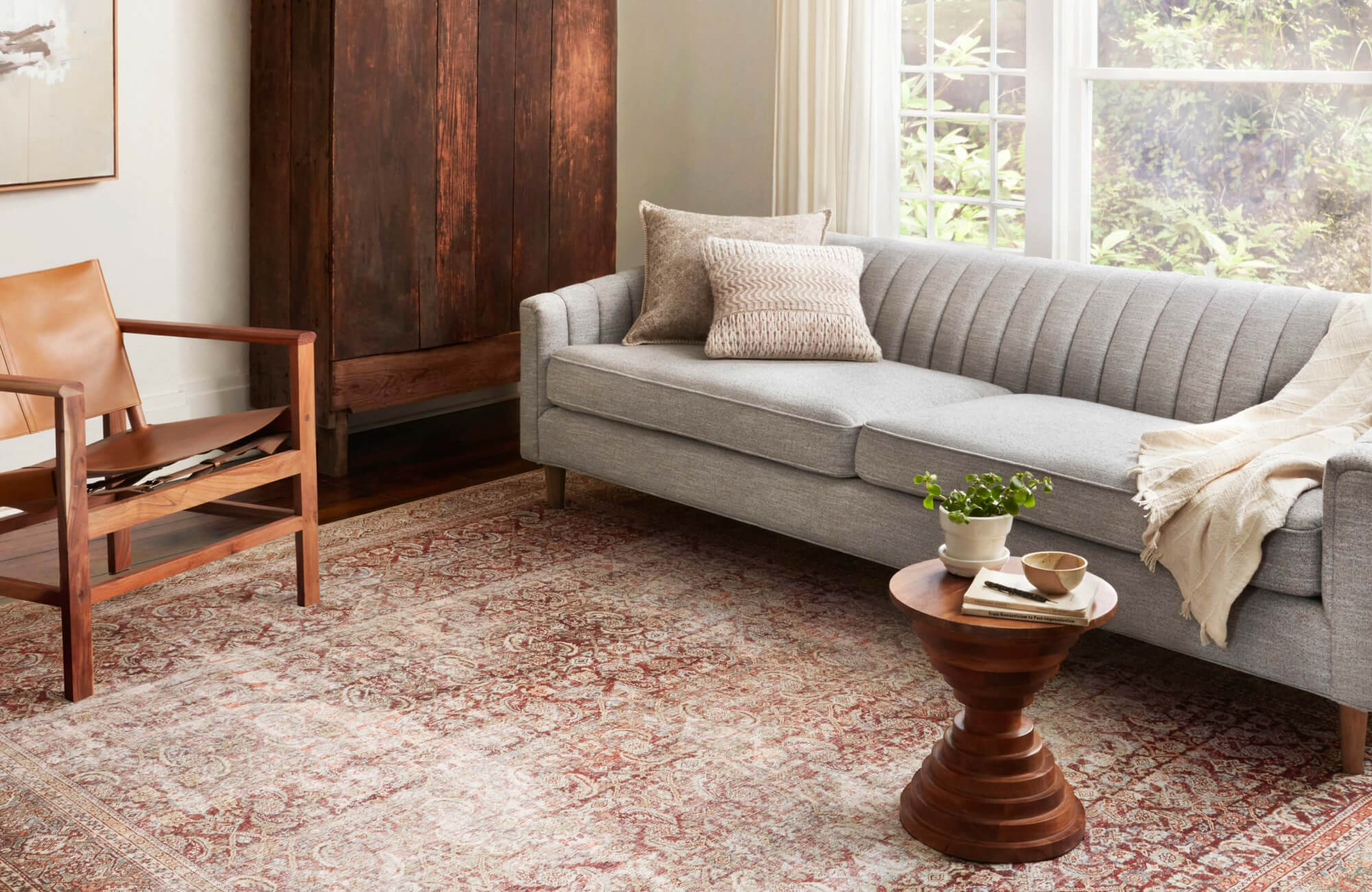 Rust and cream patterned rug in a cozy living room with a gray sofa, leather chair, wood furniture, and bright natural light from large windows.