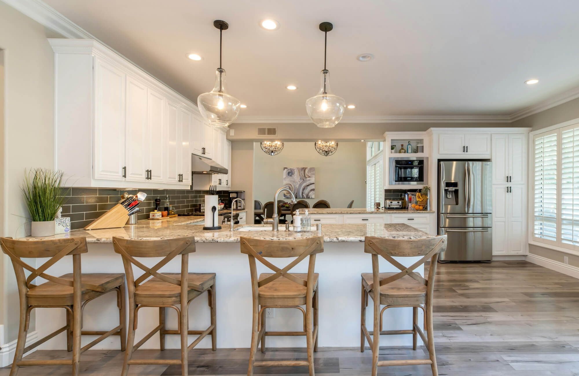 Bright, open-concept kitchen featuring white cabinetry, a granite island with wooden cross-back stools, stainless steel appliances, and two large glass pendant light fixtures that add elegant illumination over the island.