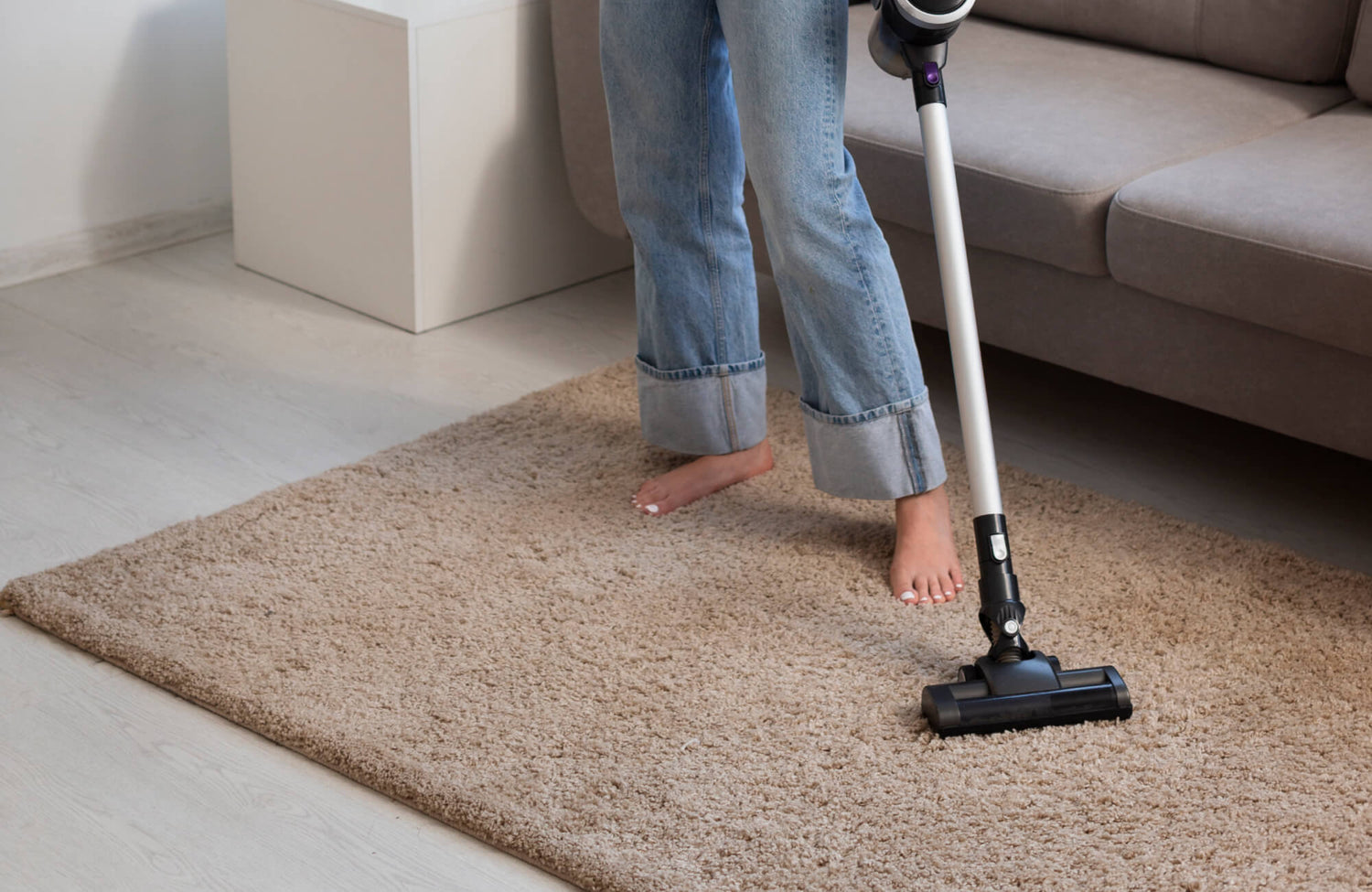 Person vacuuming a beige shag rug in a cozy living room with light wood floors and a neutral-toned sofa.