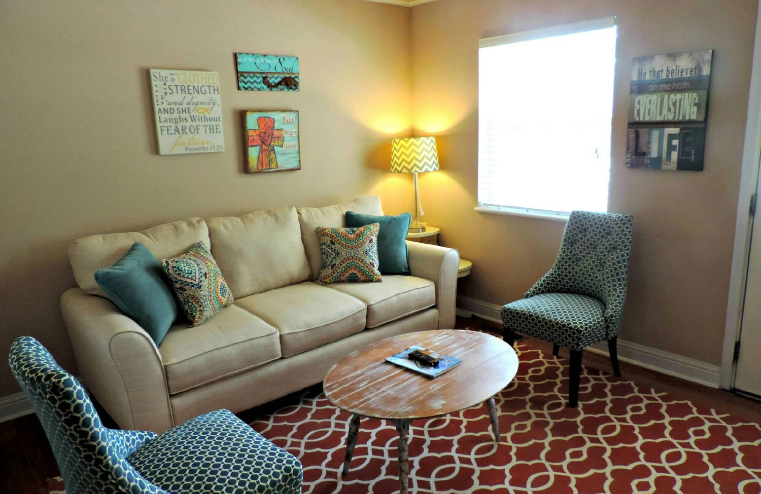 Cozy living room with a beige sofa, patterned pillows, teal accent chairs, and a bold red geometric rug.