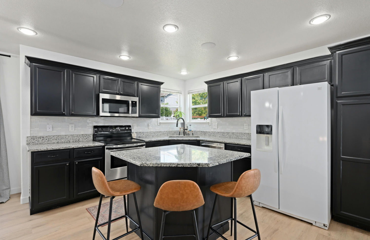 Compact kitchen with black shaker cabinets, granite island, three brown bar stools, stainless steel appliances, and recessed ceiling lights.