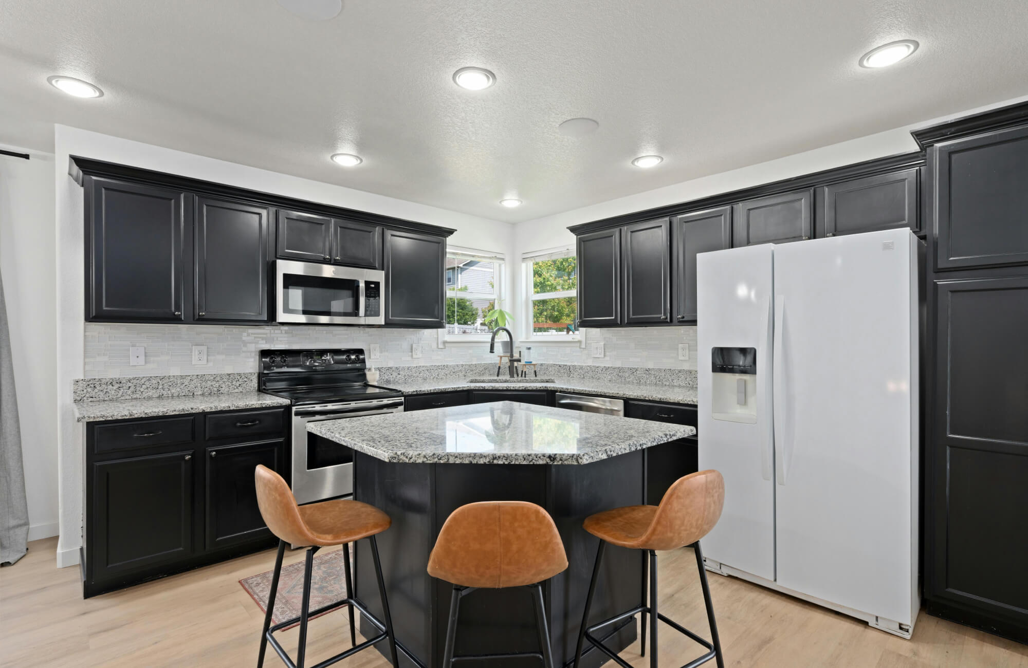 Compact kitchen with black shaker cabinets, granite island, three brown bar stools, stainless steel appliances, and recessed ceiling lights.