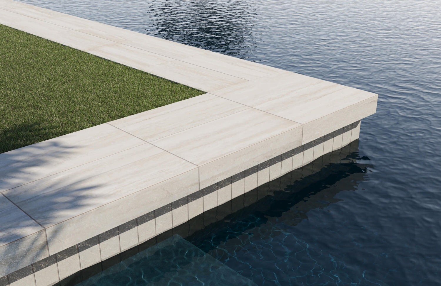 Beige porcelain travertine-look pool coping tiles with a dark waterline accent, bordered by lush green lawn and rippling blue water.