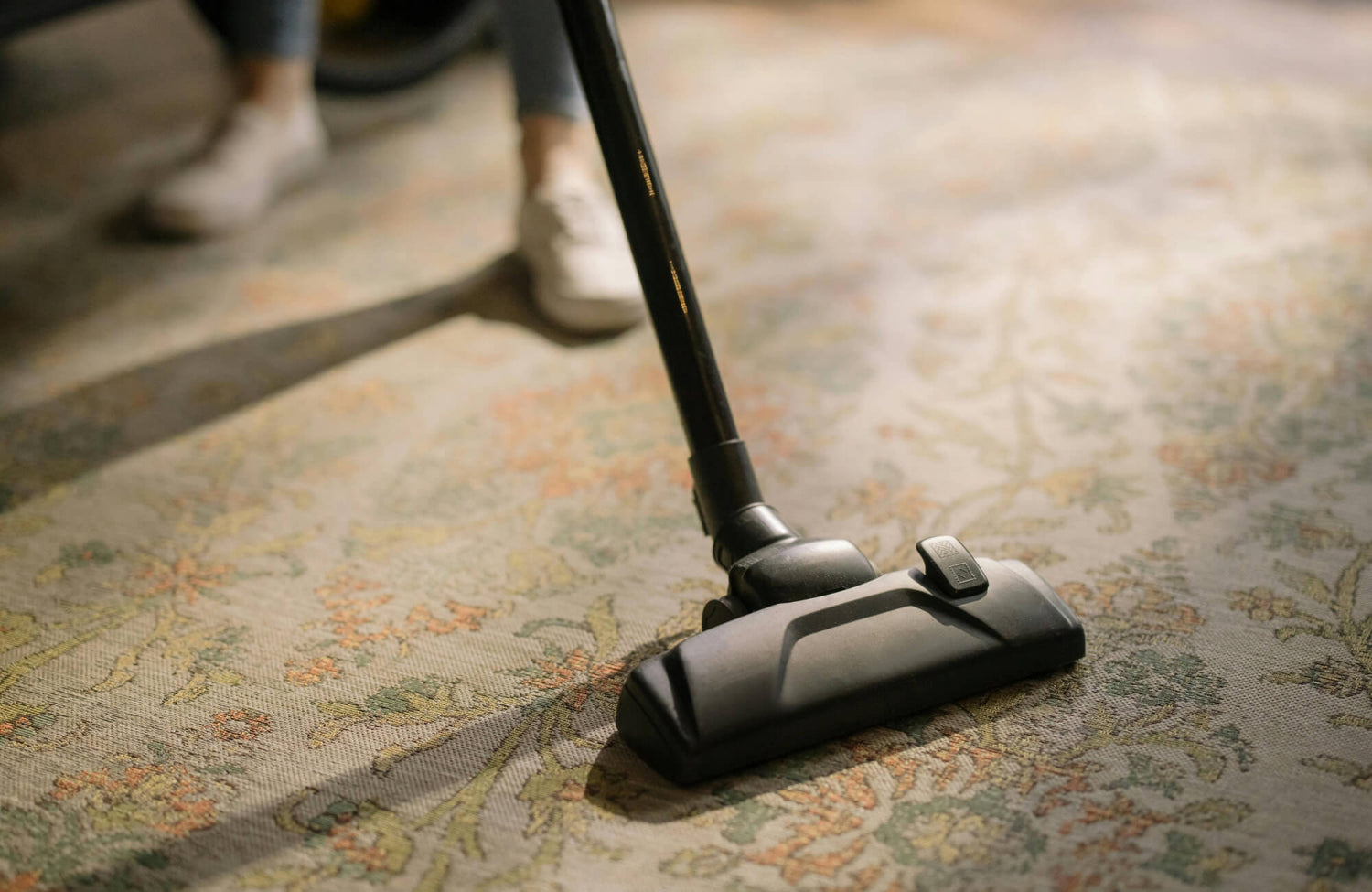 Close-up of a vacuum cleaning a vintage-style area rug with soft floral patterns in muted greens, oranges, and neutrals.