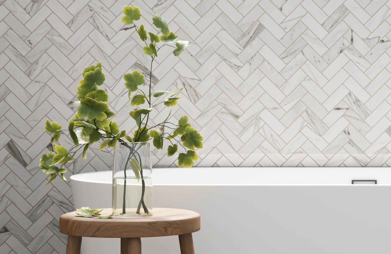 Herringbone subway tile wall behind a modern white bathtub with a wooden stool and green leafy plant in a clear glass vase.