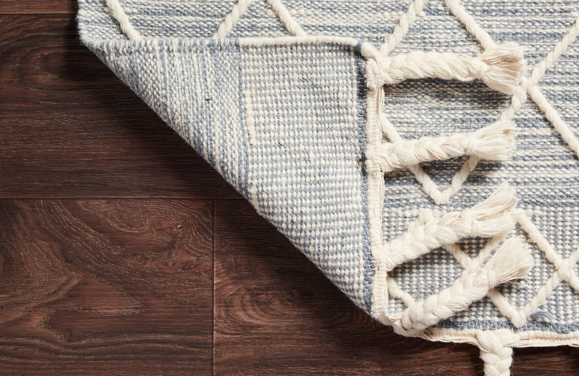 Braided cream and blue rug corner folded over dark wood flooring, revealing tightly woven backing ideal for grip and surface protection.