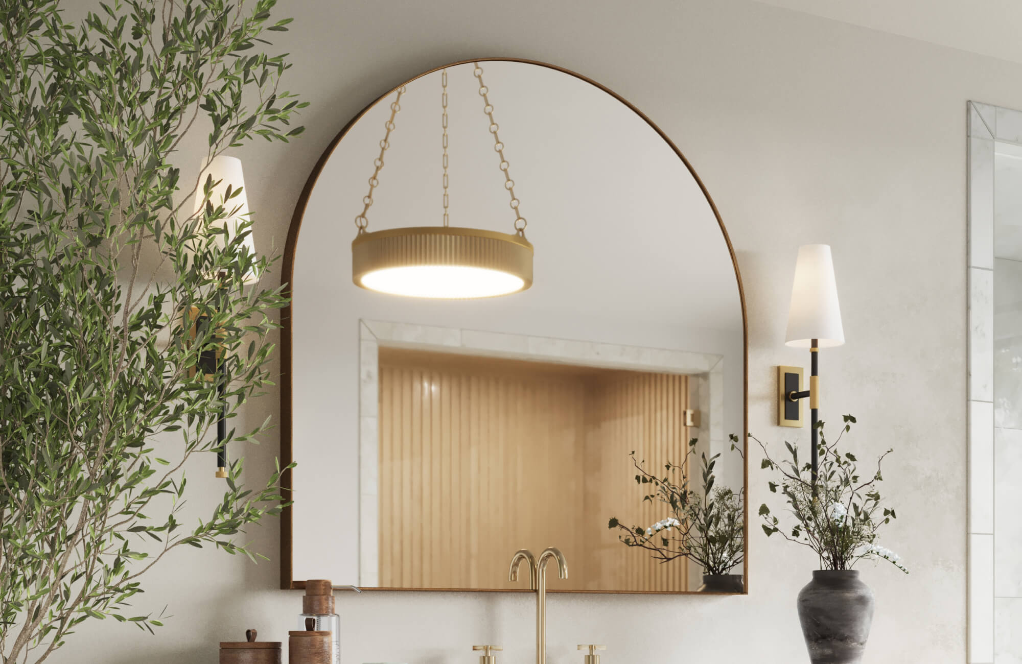 Bathroom featuring a trendy large arched mirror with thin brass frame, paired with tapered wall sconces, gold faucet, and natural greenery for a soft modern look