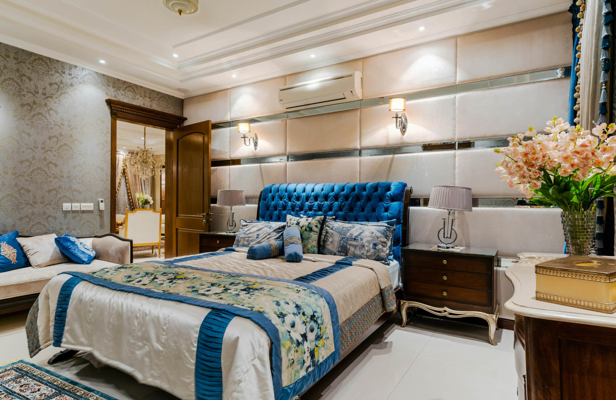 Elegant bedroom with blue tufted headboard, wall sconces, ceiling lights, floral decor, and matching patterned bedding.