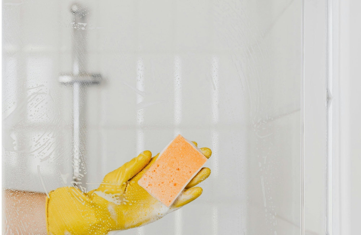 A close-up view shows someone cleaning a glass shower door with an orange sponge, revealing white shower tile blurred in the background.