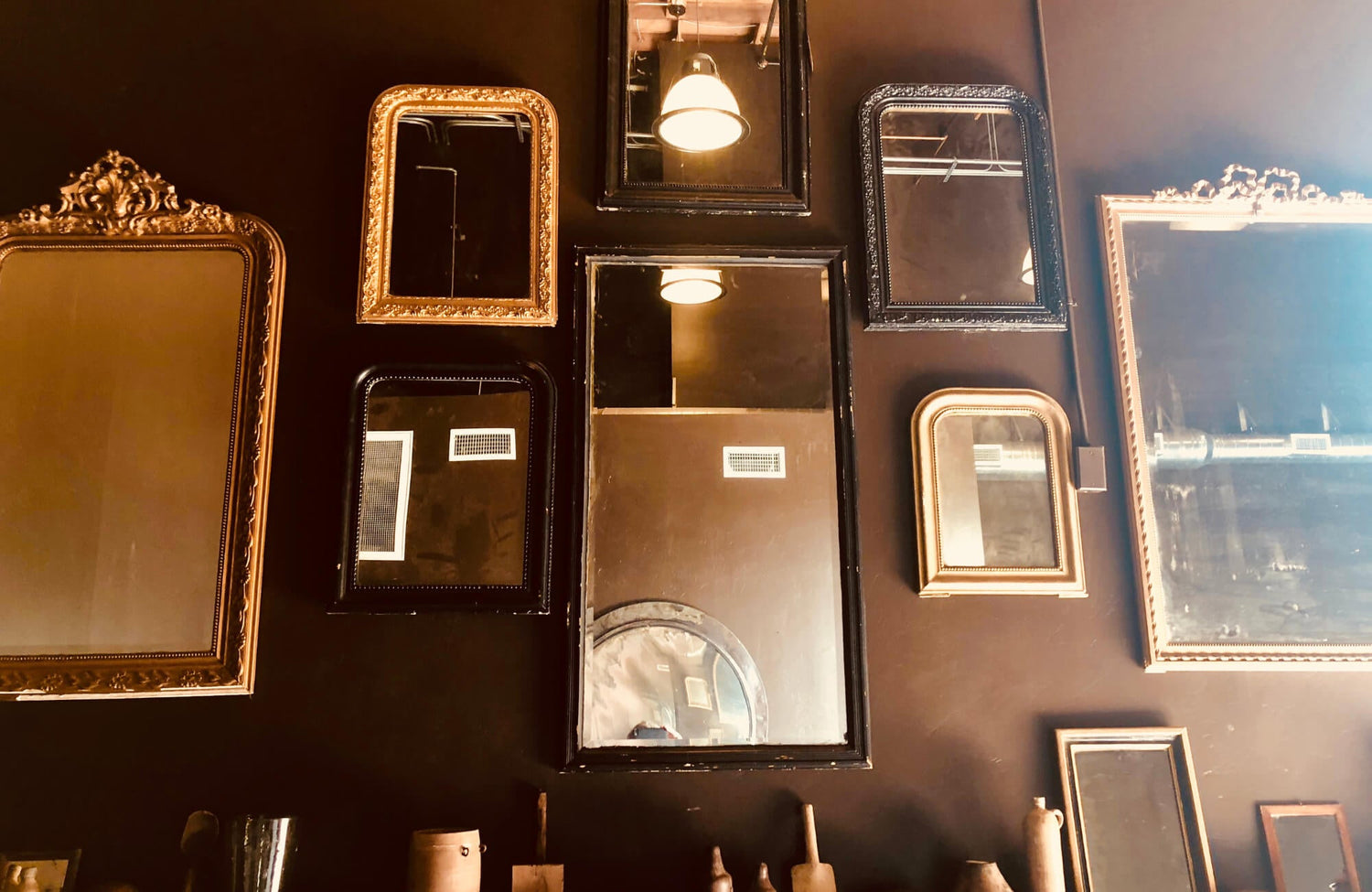 A collection of vintage brass and black-framed mirrors arranged on a dark wall, reflecting warm light and rustic decor elements.