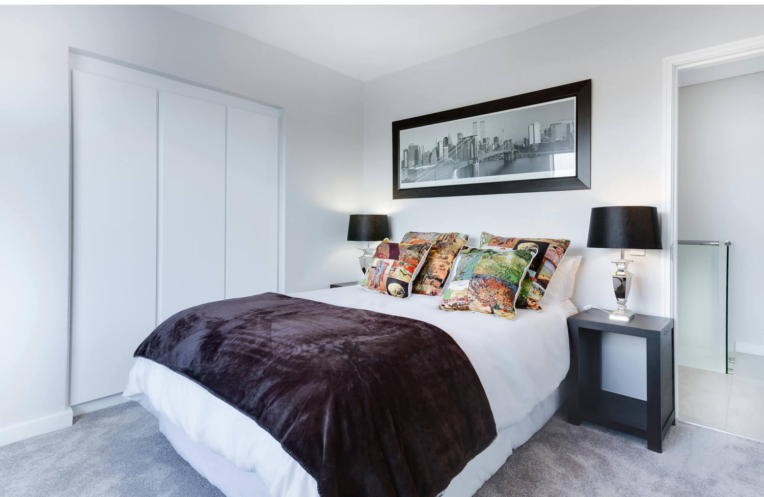 A bright, modern bedroom features a plush grey rug for bedroom comfort beneath a neatly dressed bed accented with colorful pillows, sleek black lamps, and a large monochrome cityscape artwork.