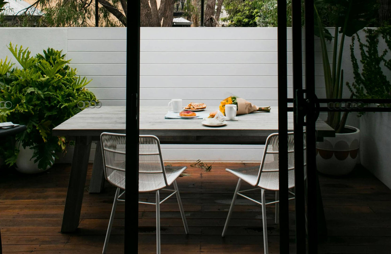 Cozy outdoor dining area with a rustic wood table, white chairs, fresh pastries, and a bouquet of sunflowers.