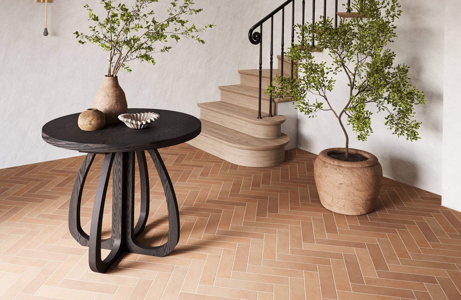 Entryway with warm terracotta herringbone tile flooring, a sculptural black table, potted greenery, and curved wood stairs creating a grounded, inviting look.