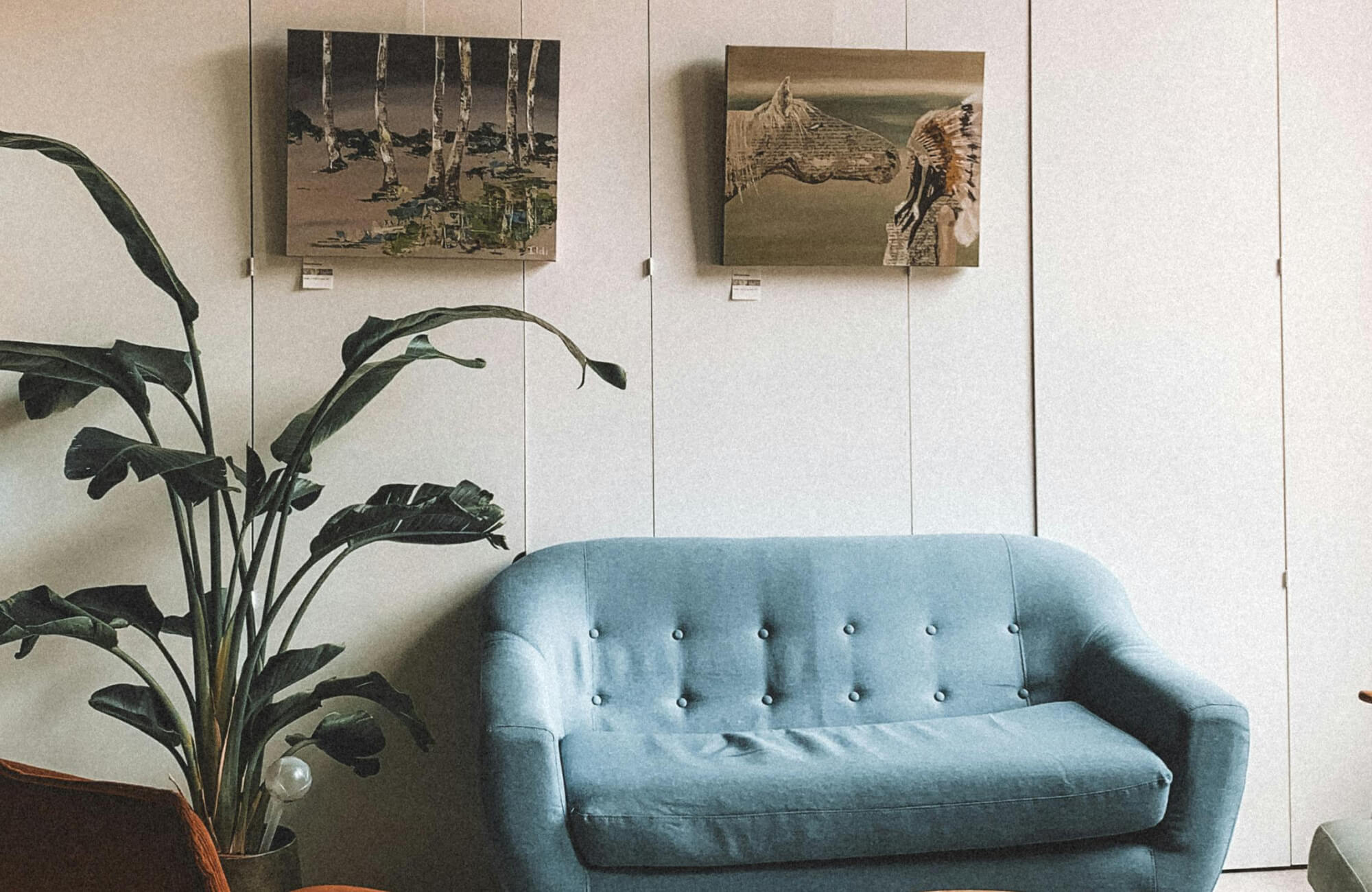 Cozy living space with a tufted blue sofa, a leafy plant, and framed wall art above a soft neutral paneled wall.