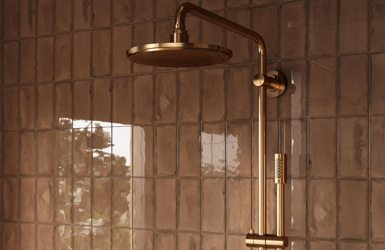 Close-up of a brass rainfall shower head mounted on a glossy brown subway tile shower wall, with vertical rectangular tiles reflecting soft window light.