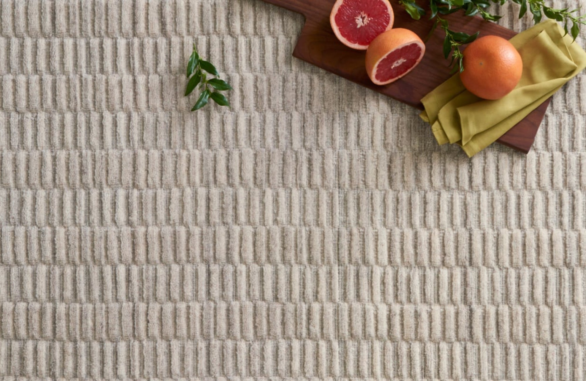 Textured beige rug with a modern ribbed pattern, styled with fresh citrus and greenery on a wooden board.