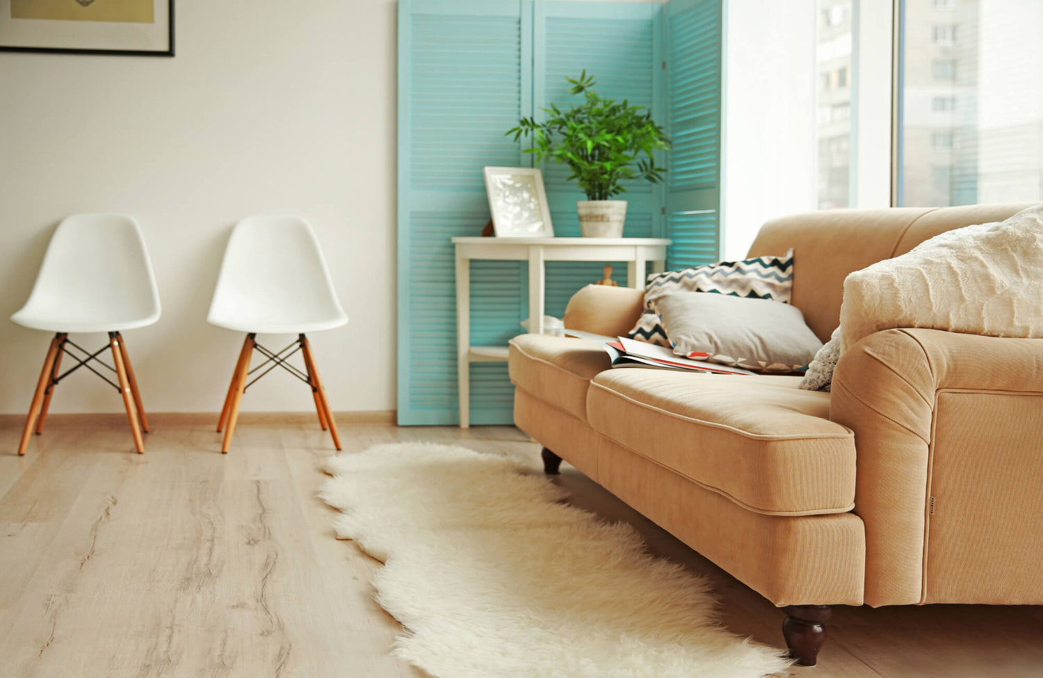 Cozy living room with a tan sofa, white wool rug on light wood tile floor, modern white chairs, and a blue folding screen with greenery.