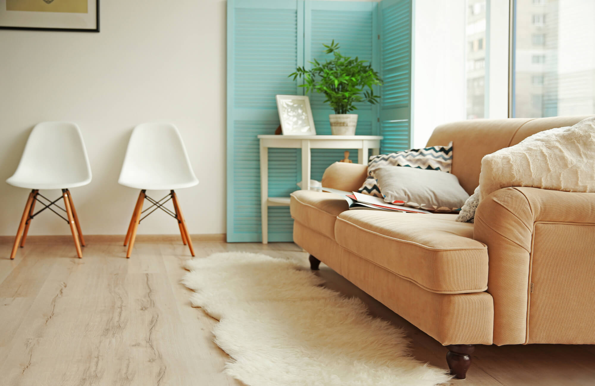 Cozy living room with a tan sofa, white wool rug on light wood tile floor, modern white chairs, and a blue folding screen with greenery.