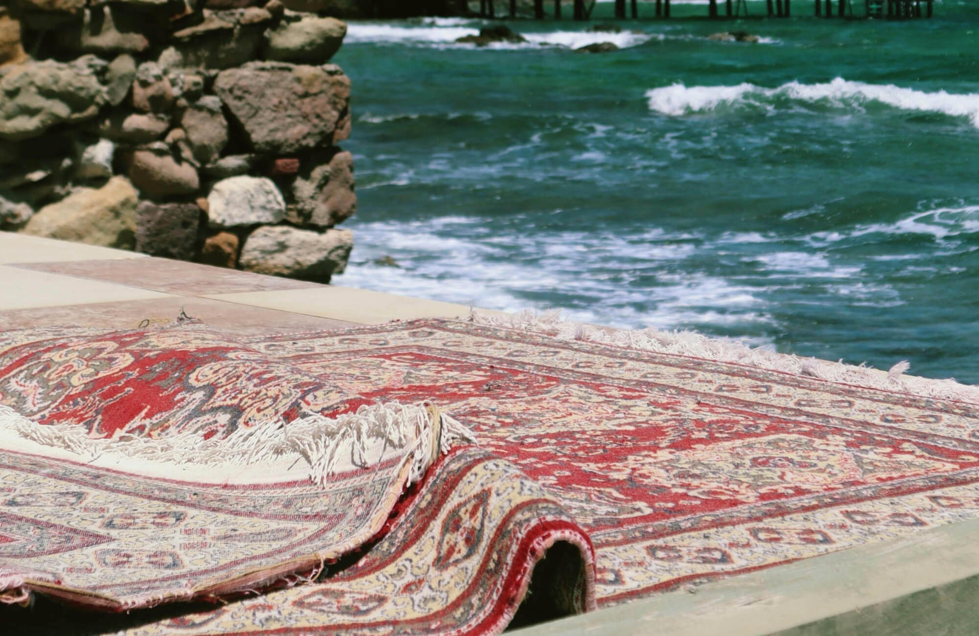 Red patterned outdoor rug with fringe edges placed near a stone wall by the ocean, highlighting durability in a coastal setting.