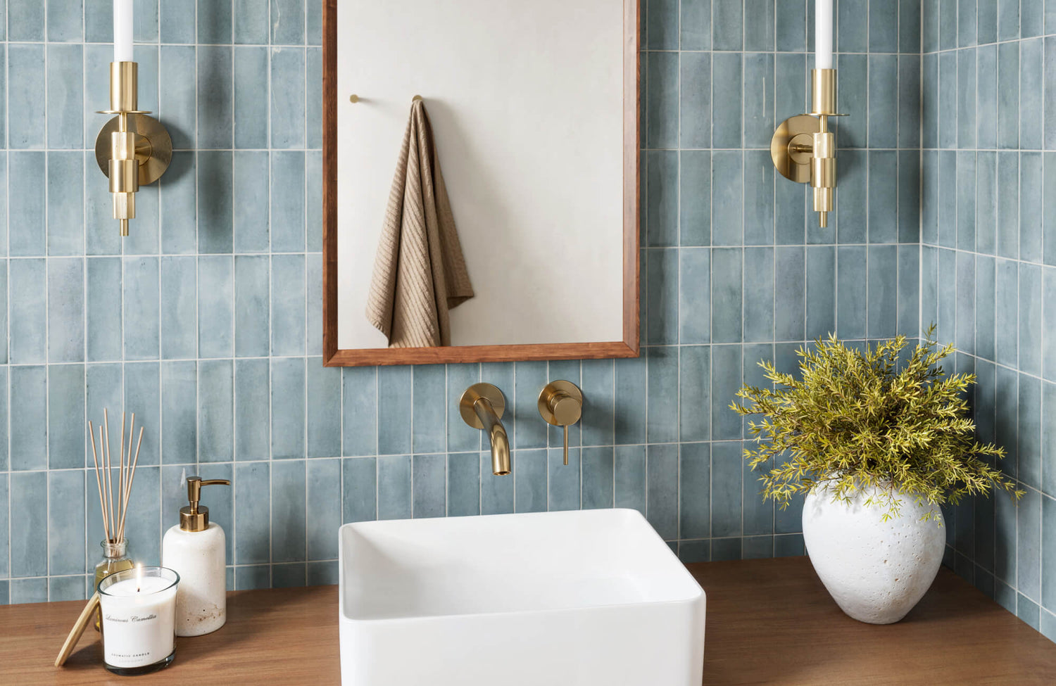 Stylish bathroom featuring vertical blue tiles, gold fixtures, a wooden vanity, and decorative accents like a potted plant and a scented candle.