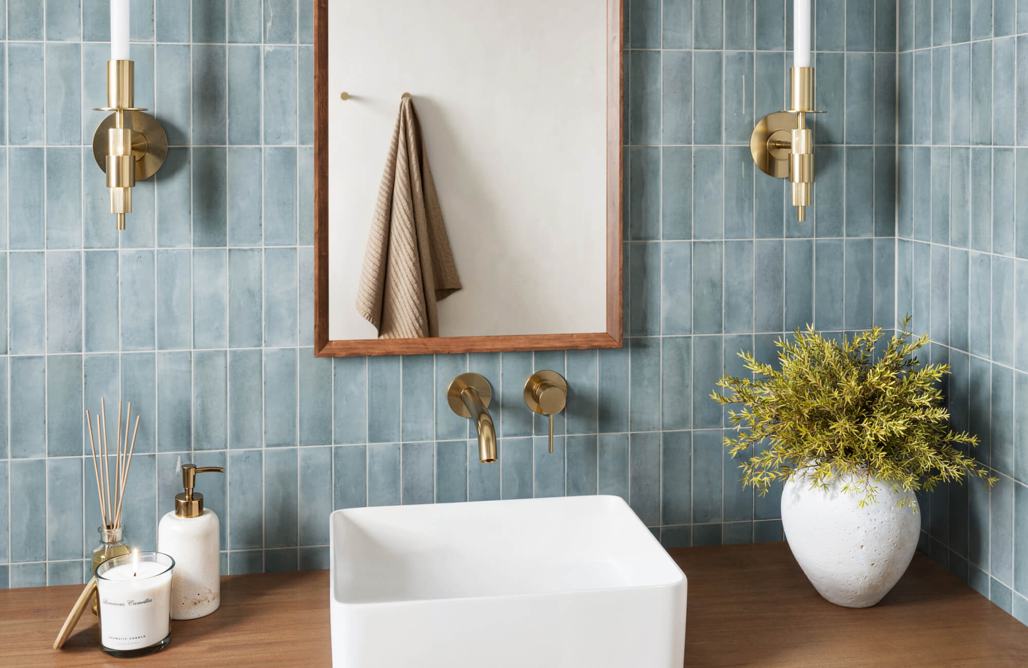 Stylish bathroom featuring vertical blue tiles, gold fixtures, a wooden vanity, and decorative accents like a potted plant and a scented candle.