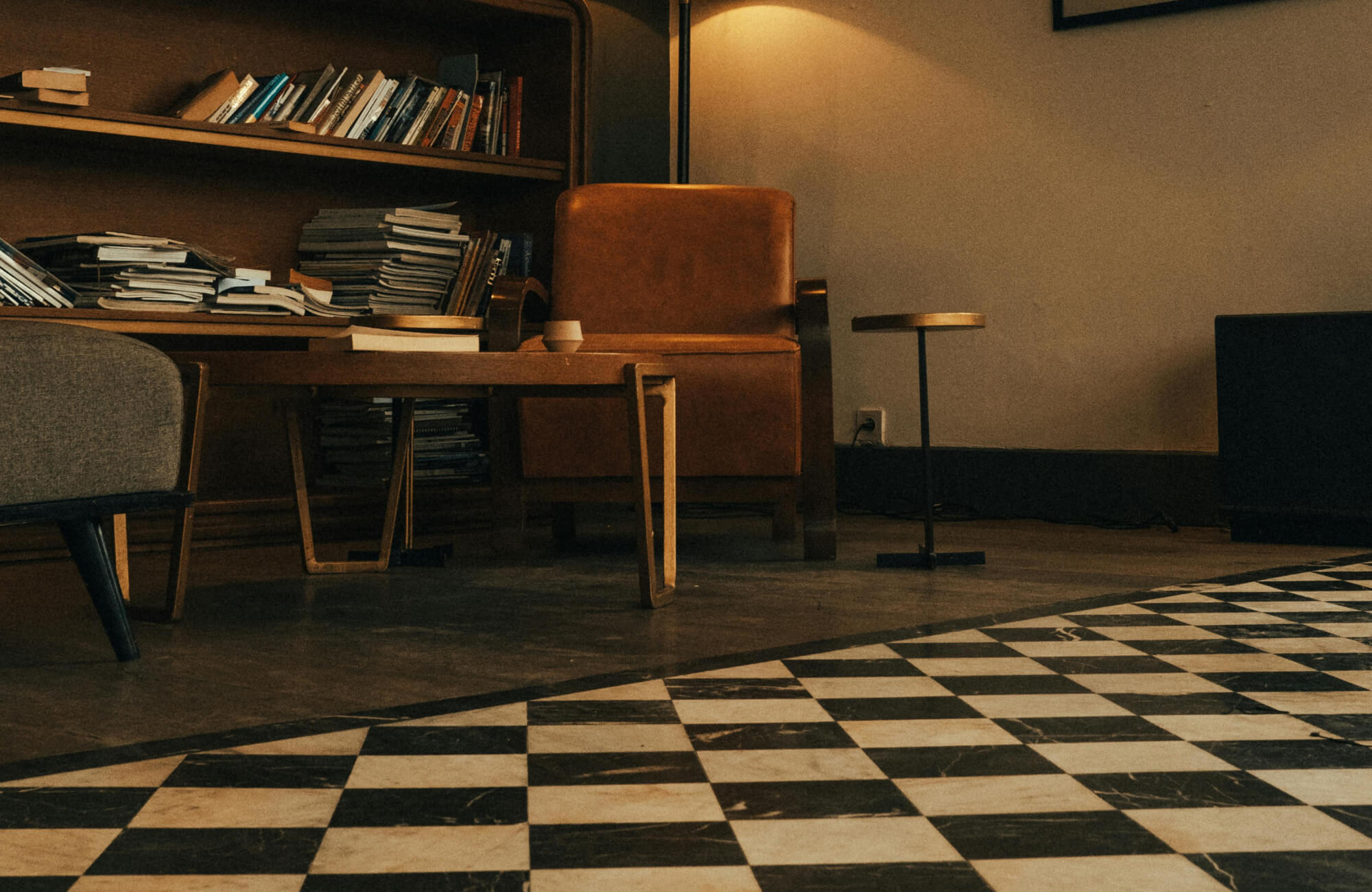 Checkerboard tile flooring adds contrast to a cozy reading nook with warm lighting, stacked books, and mid-century modern furniture.
