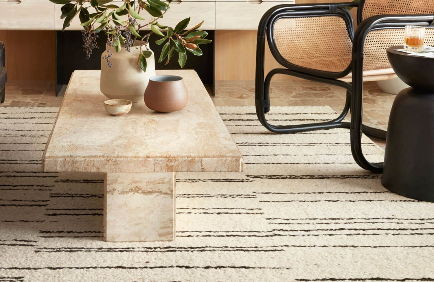 Minimalist travertine coffee table on cream striped rug, paired with modern cane chairs and ceramic decor.