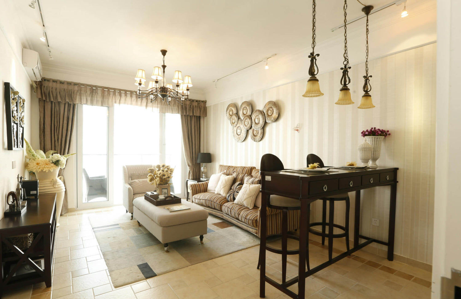 Warm beige living room featuring a striped sofa, chandelier, tile flooring, and a neutral area rug with soft geometric accents.