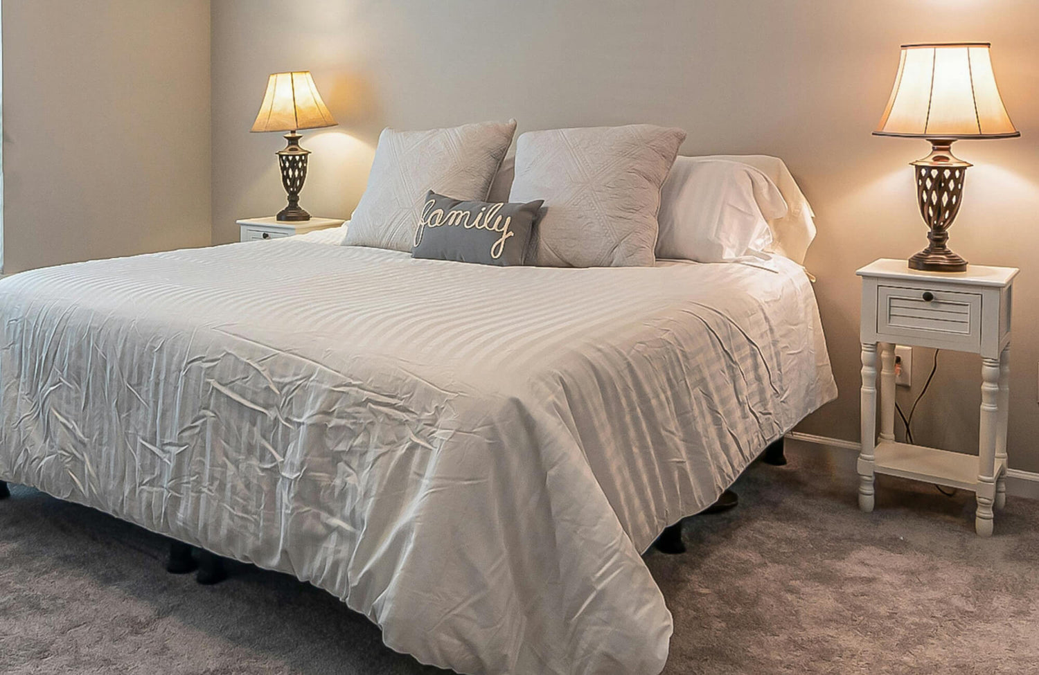 A cozy bedroom featuring a neatly made queen bed with crisp white bedding, soft neutral pillows, matching bedside tables, and warm ambient lighting from two elegant lamps.
