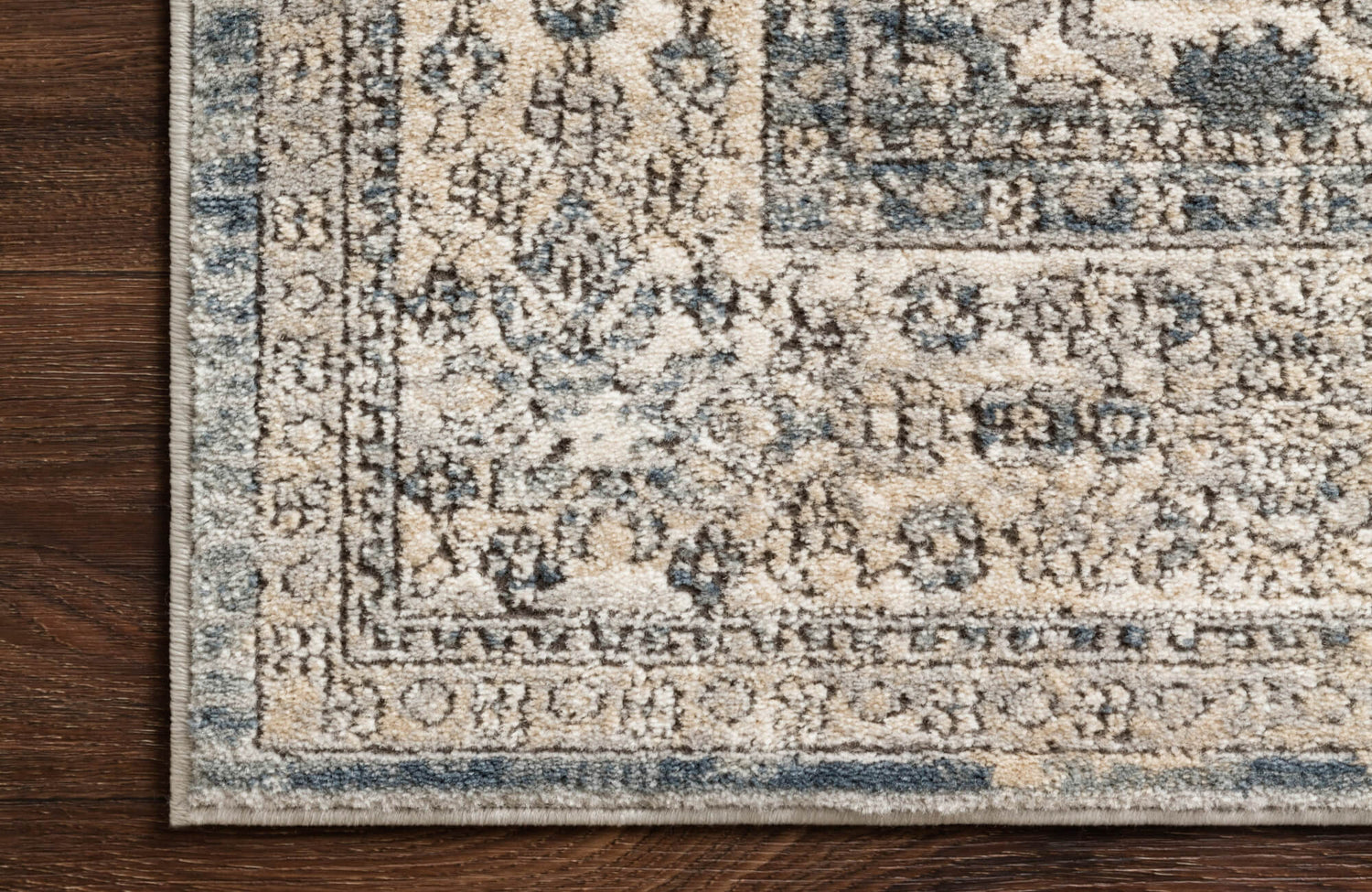 Close-up of a polypropylene rug corner featuring a classic floral border in muted blue and beige tones atop rich wood flooring.