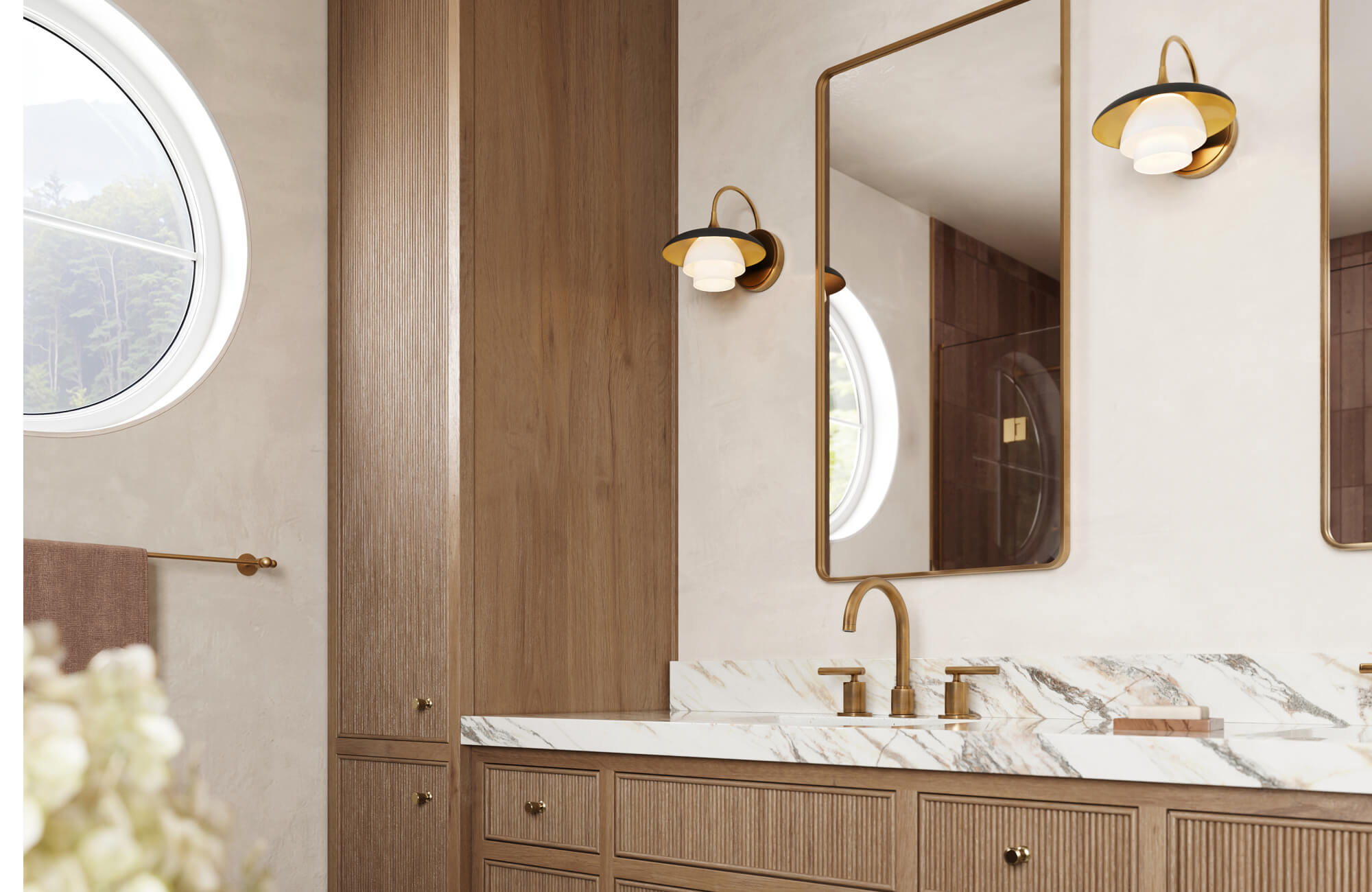 A warm contemporary bathroom featuring a sculptural brass wall light with a tiered opal glass shade, illuminating wood cabinetry, marble countertops, and large framed mirrors with a soft, elegant glow.