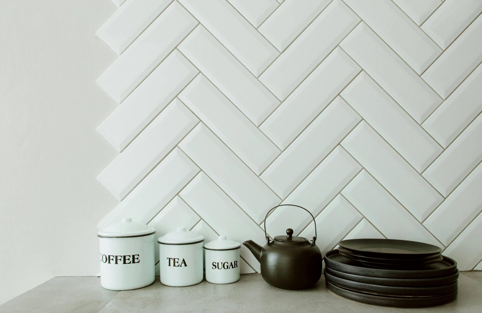 White herringbone tile backsplash with coffee, tea, and sugar jars beside a matte black teapot and stacked plates on a concrete countertop.