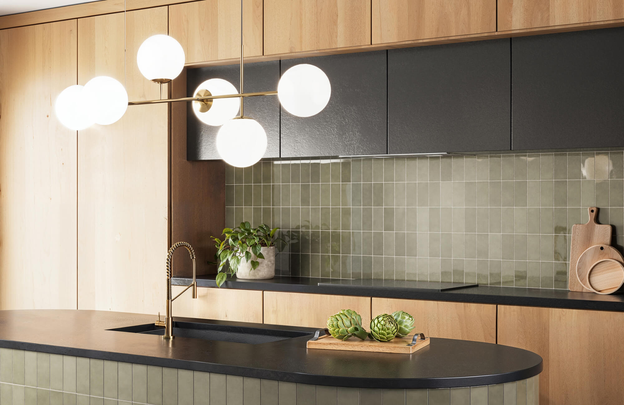 Modern kitchen with glossy olive green tile backsplash, globe pendant light, and matte black countertop over warm wood cabinetry.