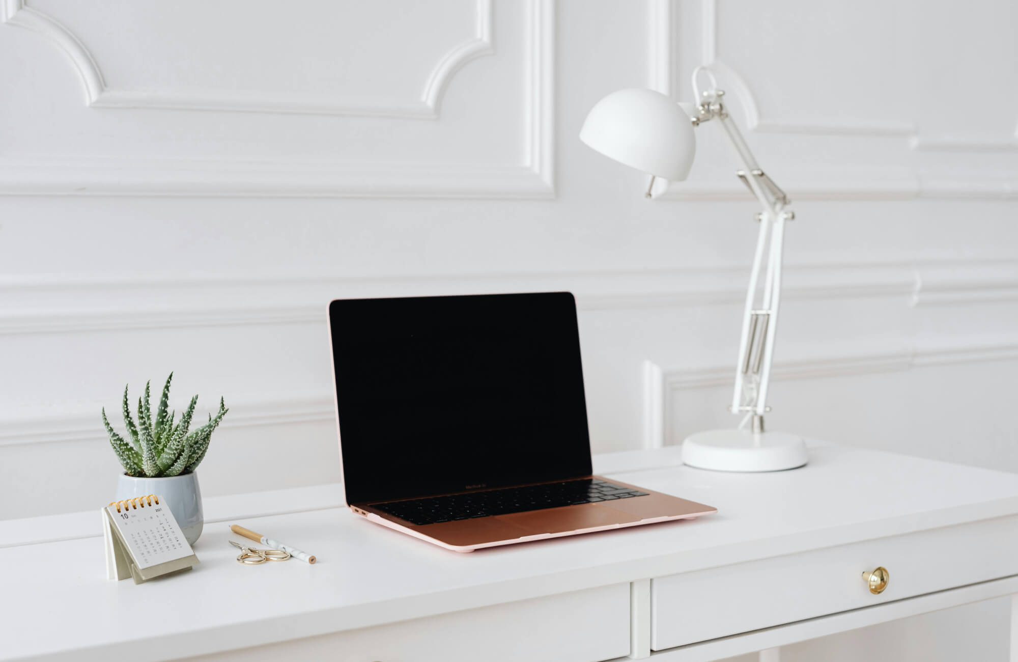 Minimalist white desk setup featuring a rose gold laptop, white task lamp, mini calendar, and a small potted plant against a white panel wall.