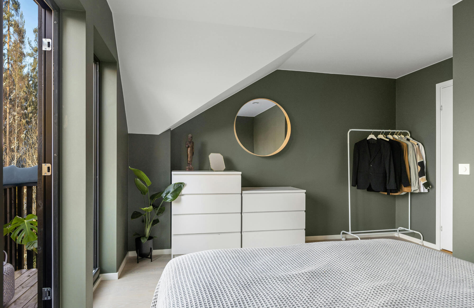 Bedroom with olive green walls, a round brass mirror above white dressers, indoor plants, and natural light from a balcony door.