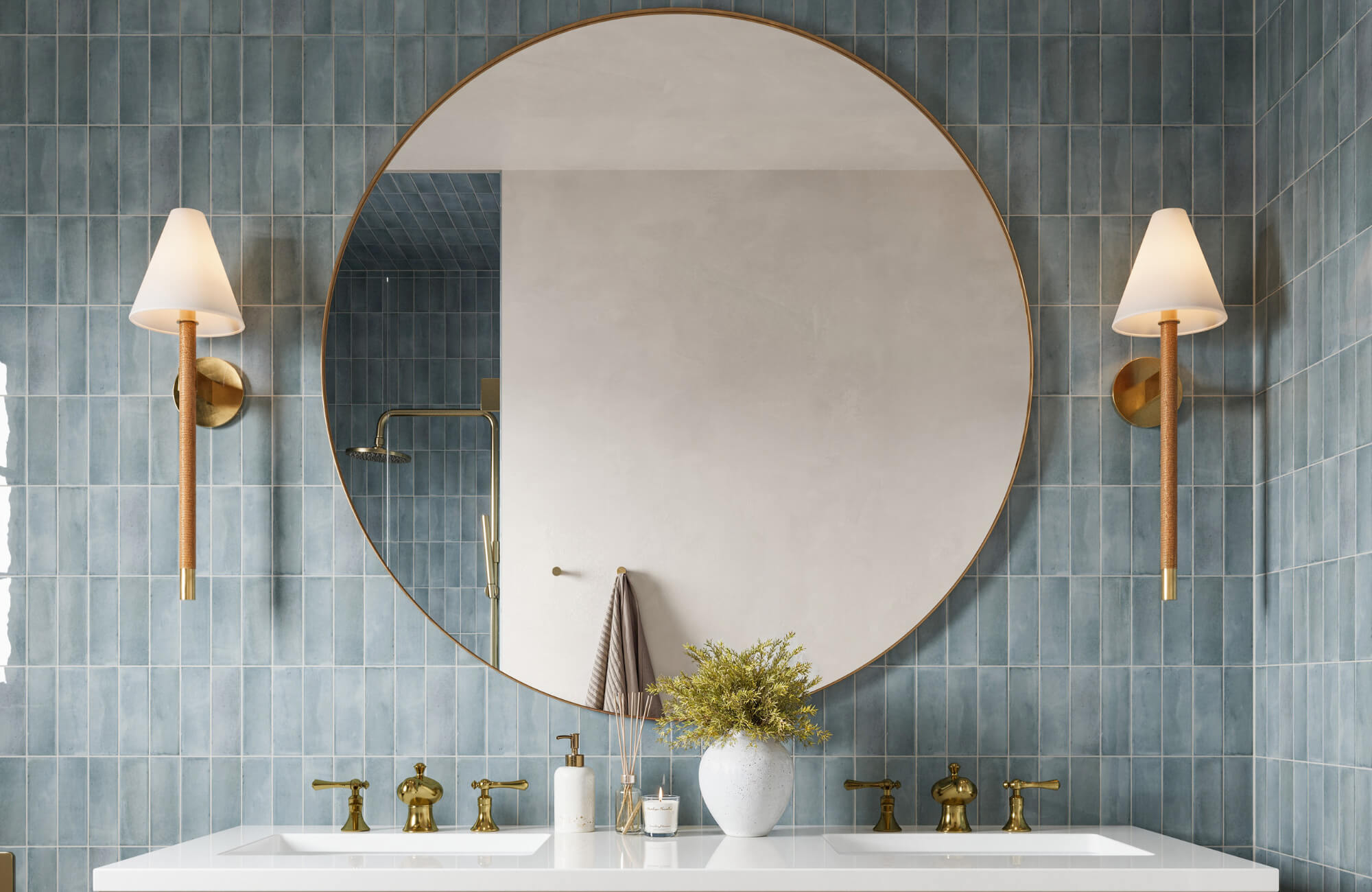Round mirror with a thin brass frame above a double vanity, flanked by wall sconces, set against vertical blue ceramic tiles.