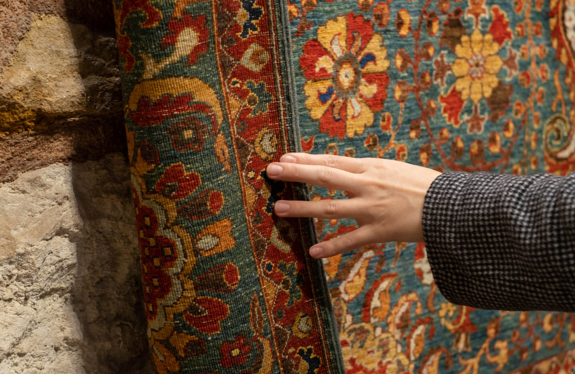 A hand brushing the edge of a rolled Persian-style rug with dense floral motifs in red, gold, and teal, set against a rough stone wall.