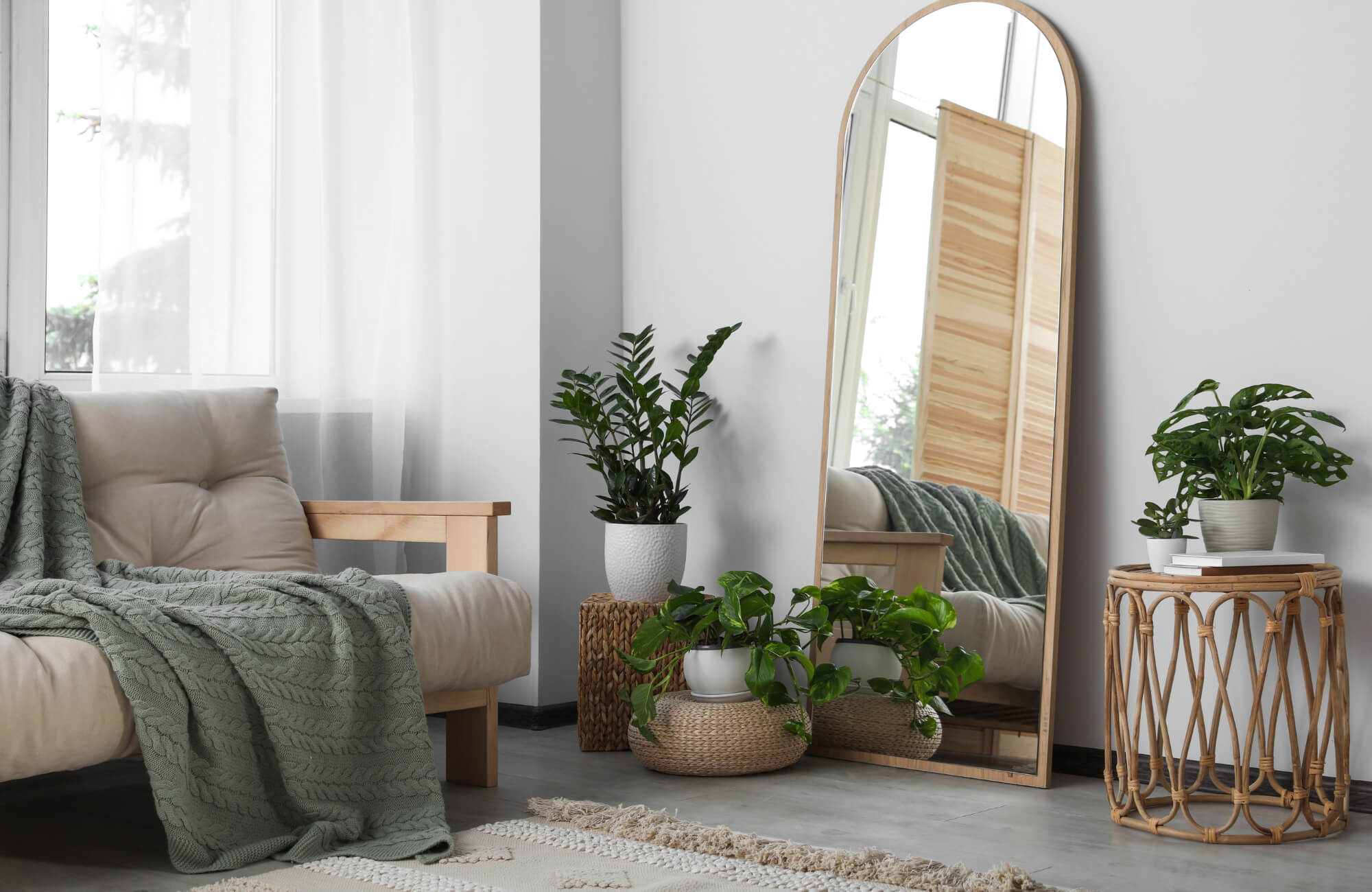 Cozy living room with a full-length arched mirror, indoor plants, and a neutral-toned sofa draped with a green knit throw blanket.