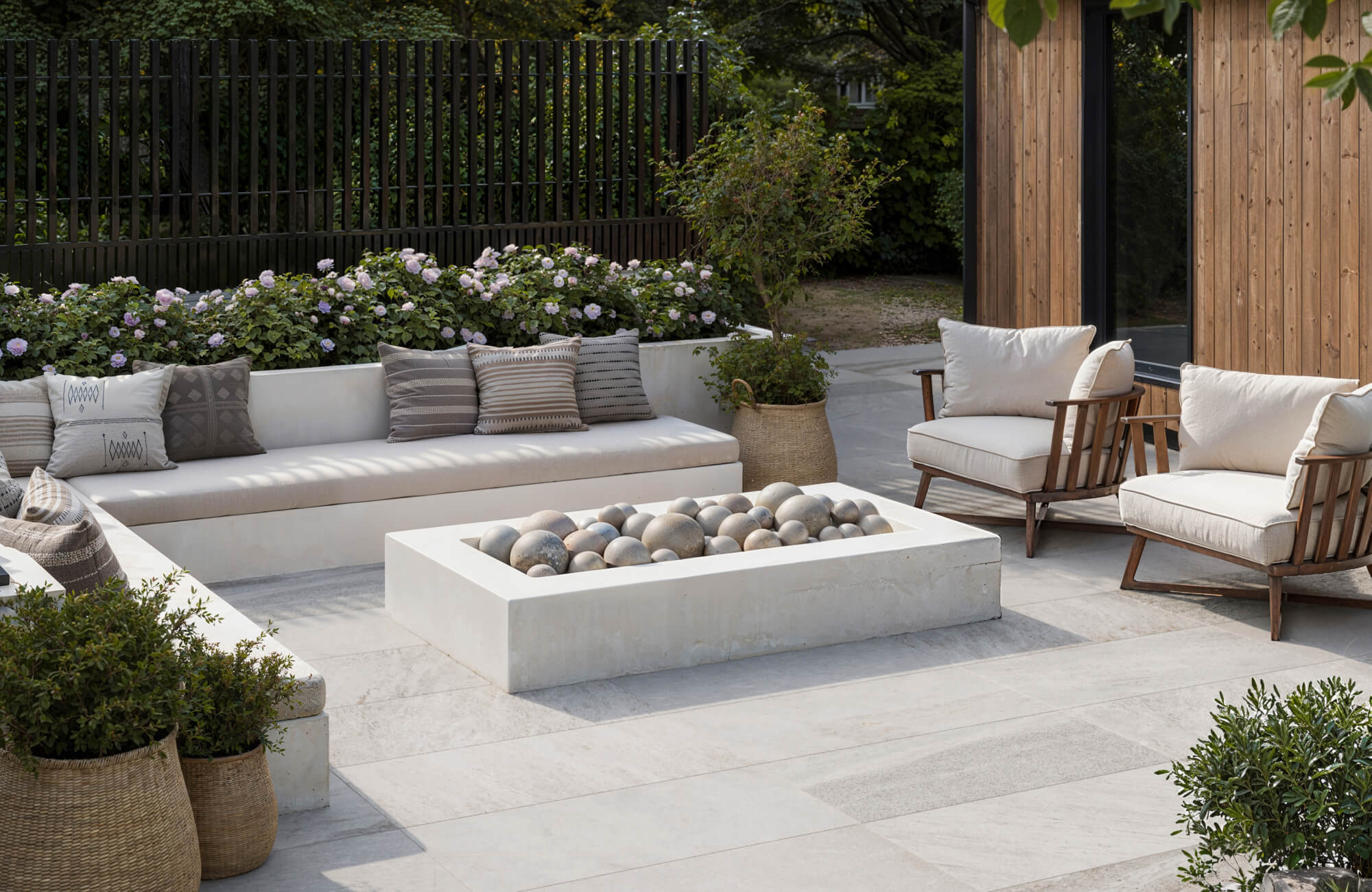 Modern outdoor seating area with porcelain tile flooring, cushioned benches, and a decorative stone fire feature surrounded by greenery.