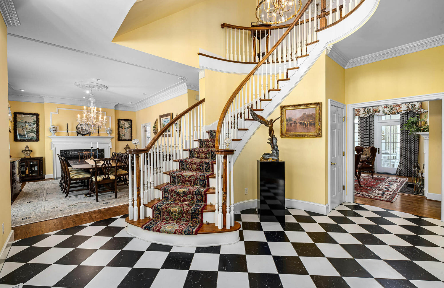 Elegant foyer with a grand curved staircase, yellow walls, and black-and-white checkerboard marble tile flooring in a diamond pattern.