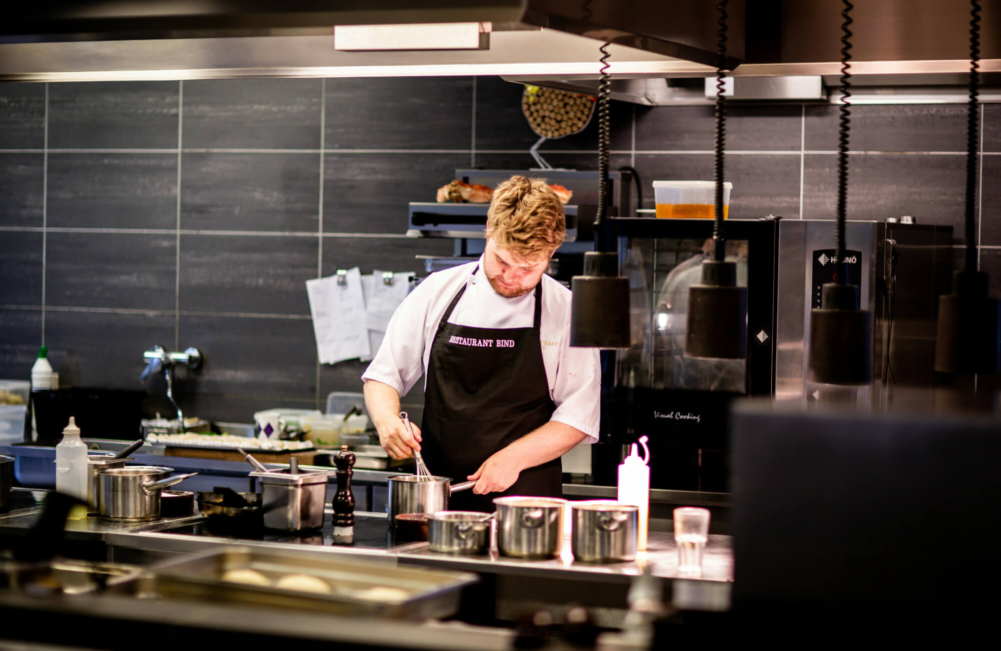 Professional chef preparing food in a modern commercial kitchen with stainless steel cookware and dark tiled walls.
