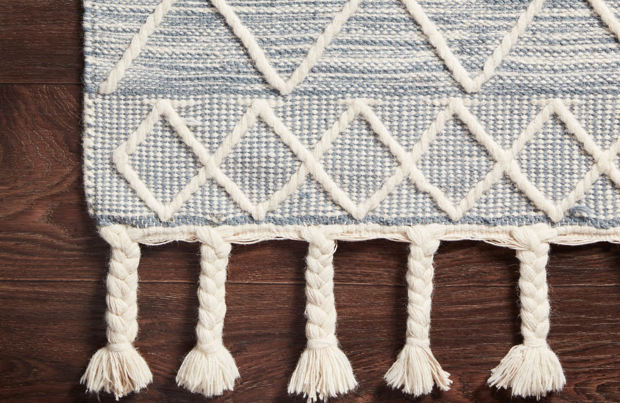 Textured rug in blue and ivory with raised diamond motifs and thick braided fringe, resting on hardwood flooring.