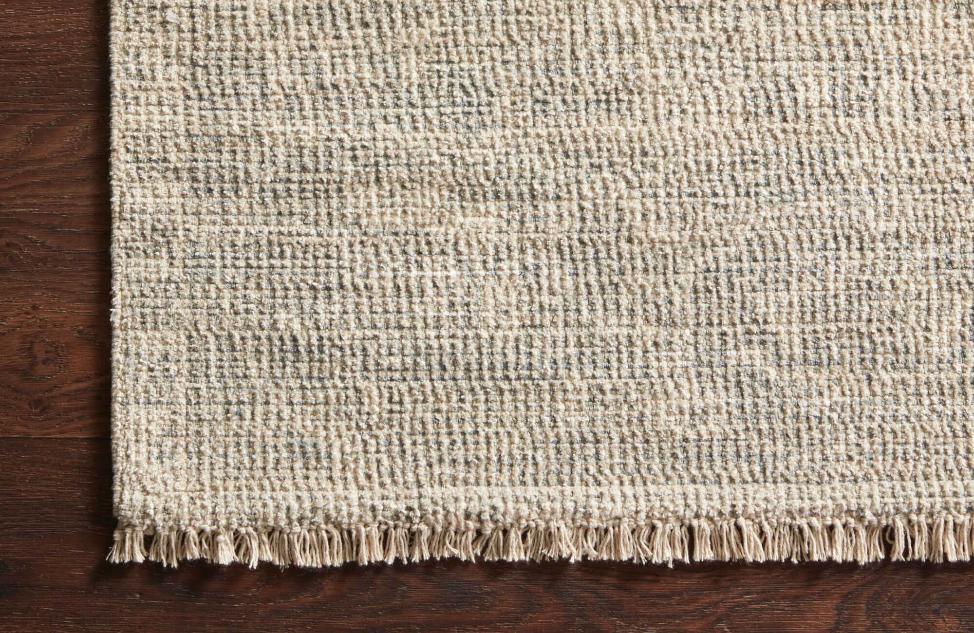 Textured flatweave rug in neutral tones with delicate fringe detailing, placed over a rich brown hardwood surface.