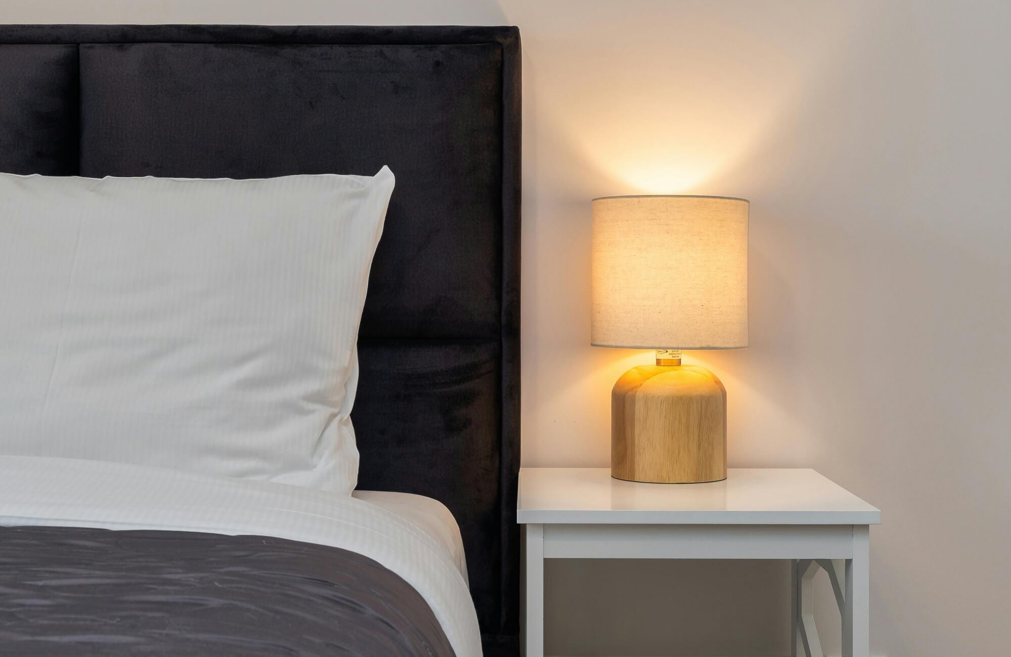 Warm bedside lamp on a white nightstand beside a neatly made bed with white linens and a dark headboard.