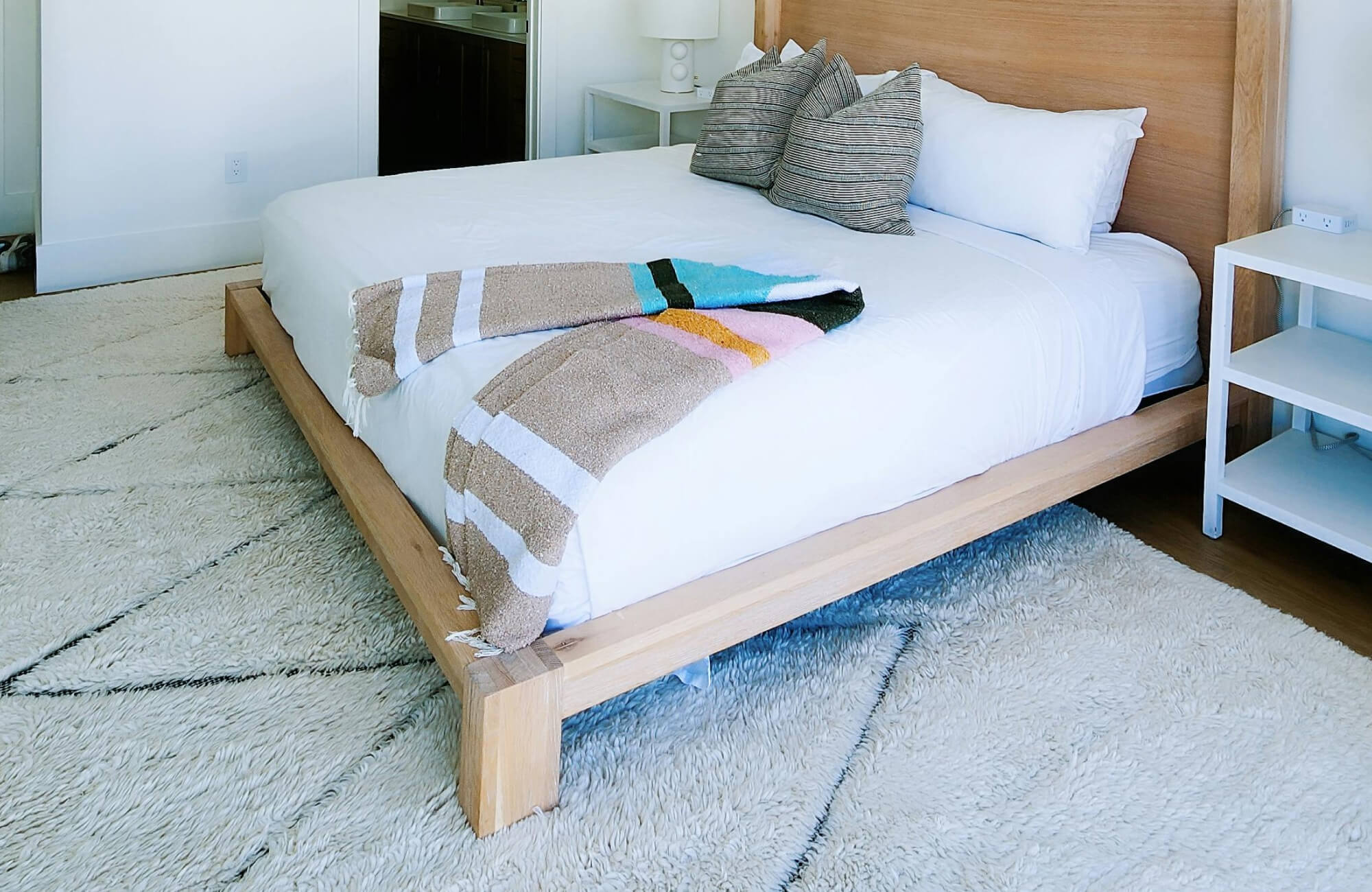 Cozy bedroom with a low-profile wooden bed frame, white bedding, striped pillows, and a soft, square geometric rug.