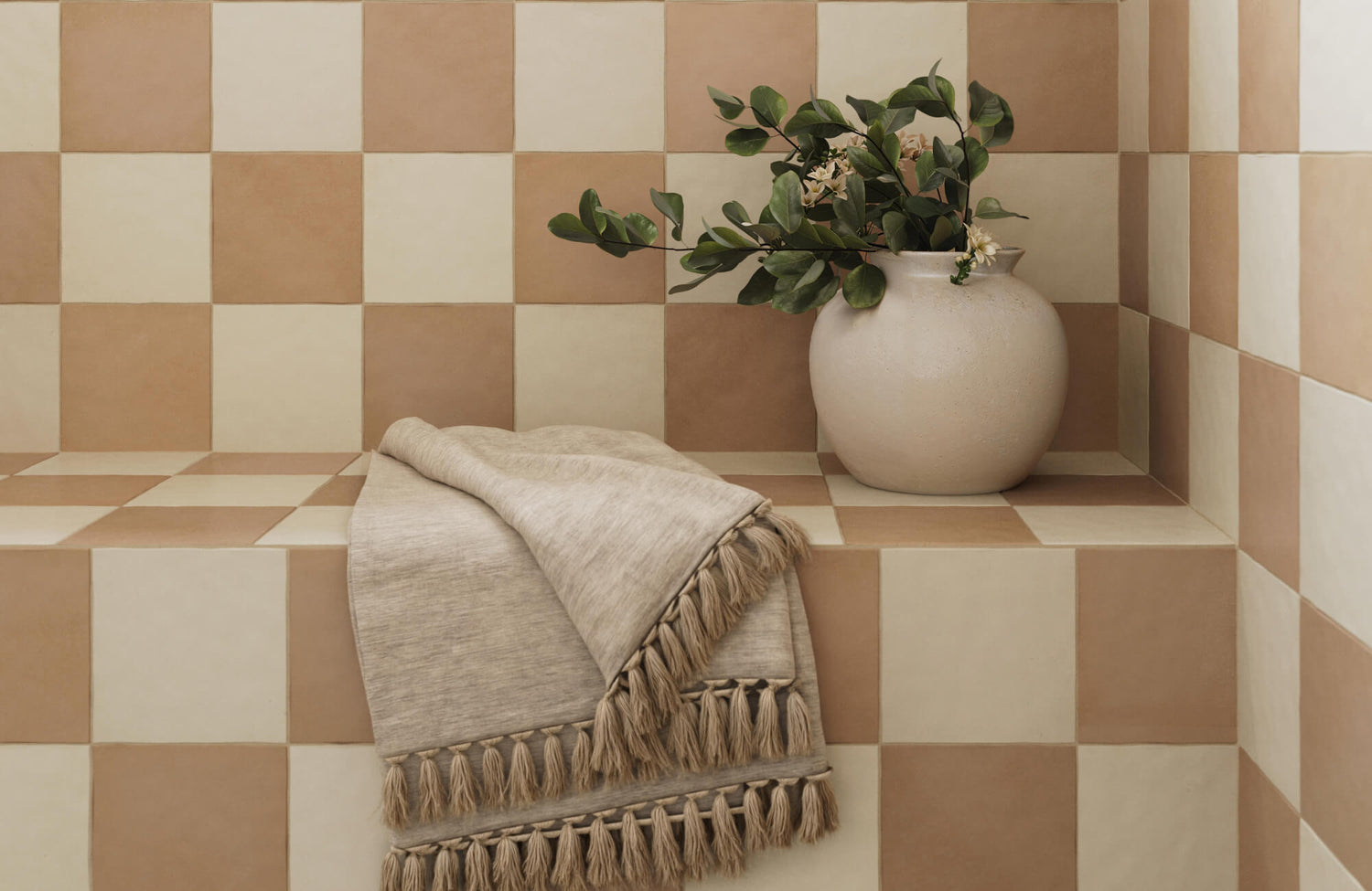 Cozy nook with terracotta and cream checkerboard tiles, a tasseled linen towel, and a ceramic vase with greenery for a warm, rustic feel.